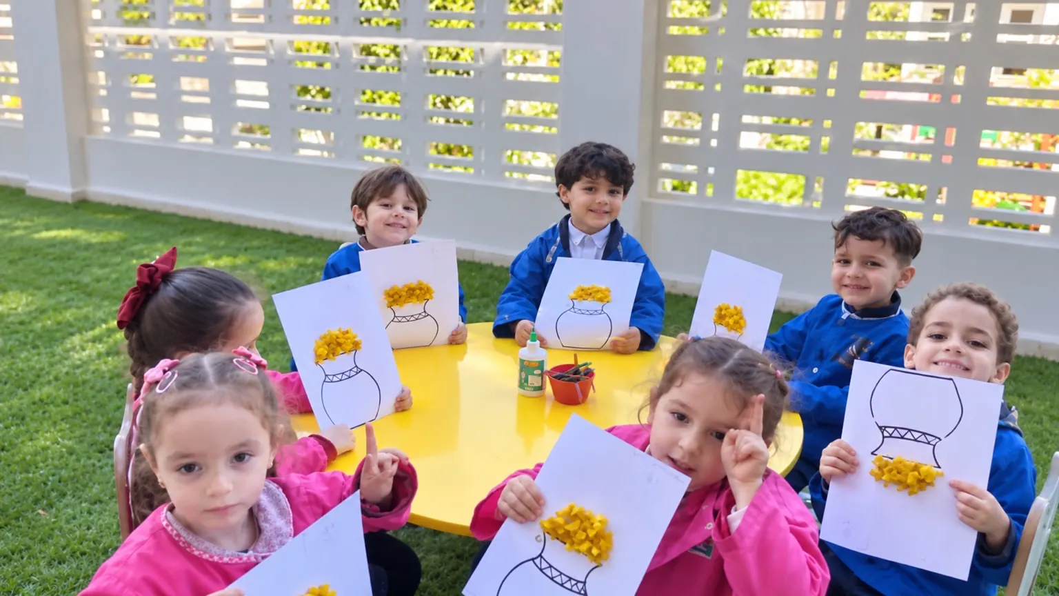 Enfants réunis autour d’une table jaune dans le jardin, présentant leurs activités manuelles